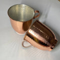 Two hammered copper mugs with a silver-toned interior, shown on a light gray background with one standing upright and the other lying on its side.