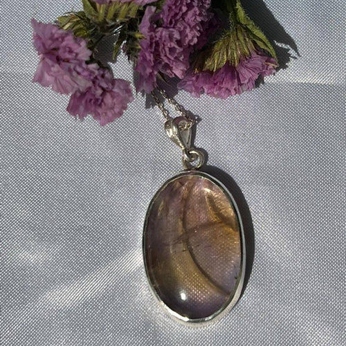 Necklace with a large oval pendant and chain, displayed with purple flowers on a textured surface.