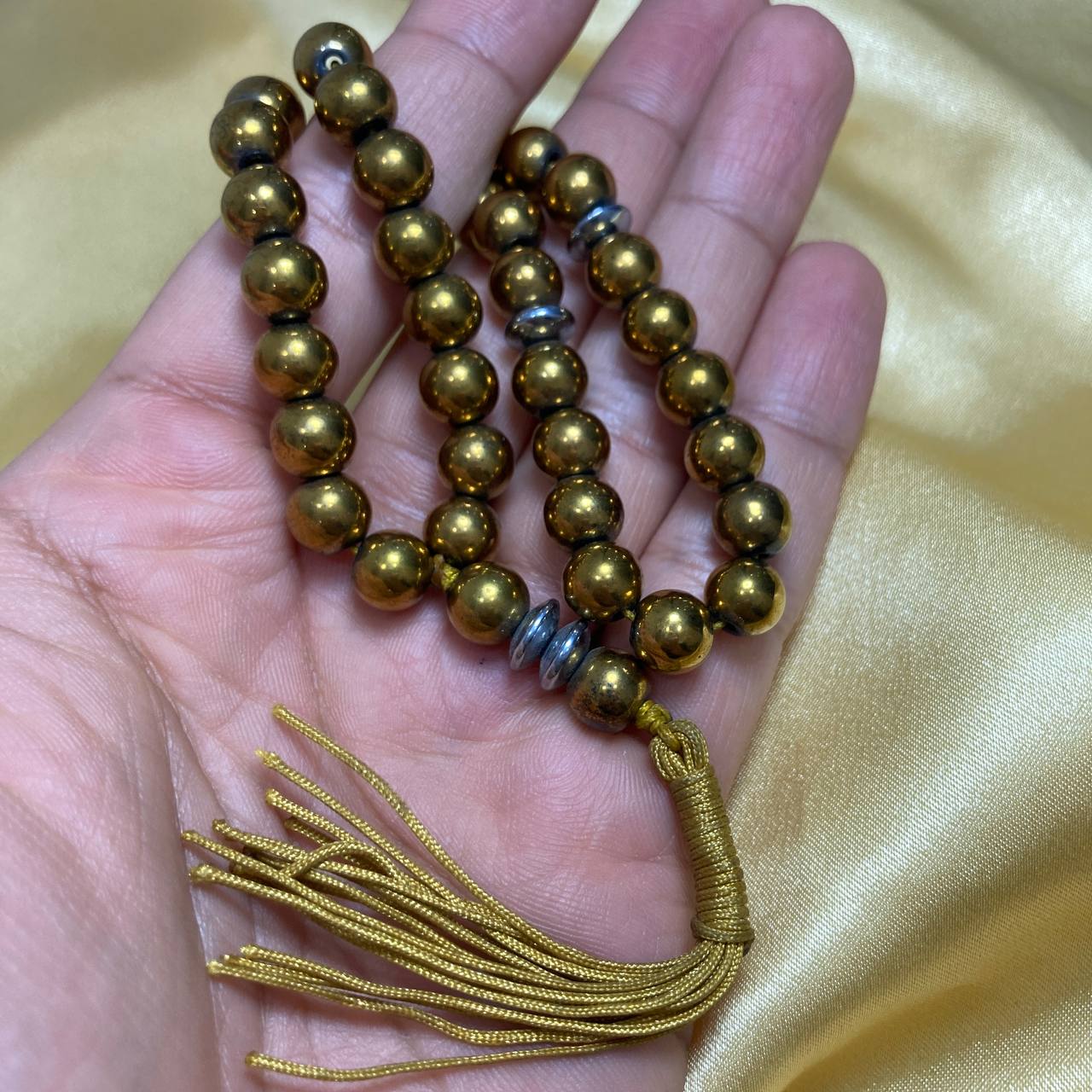 Gold beaded necklace with tassel held in a hand against a gold fabric background Golden Hematite 33 Beads Rosary