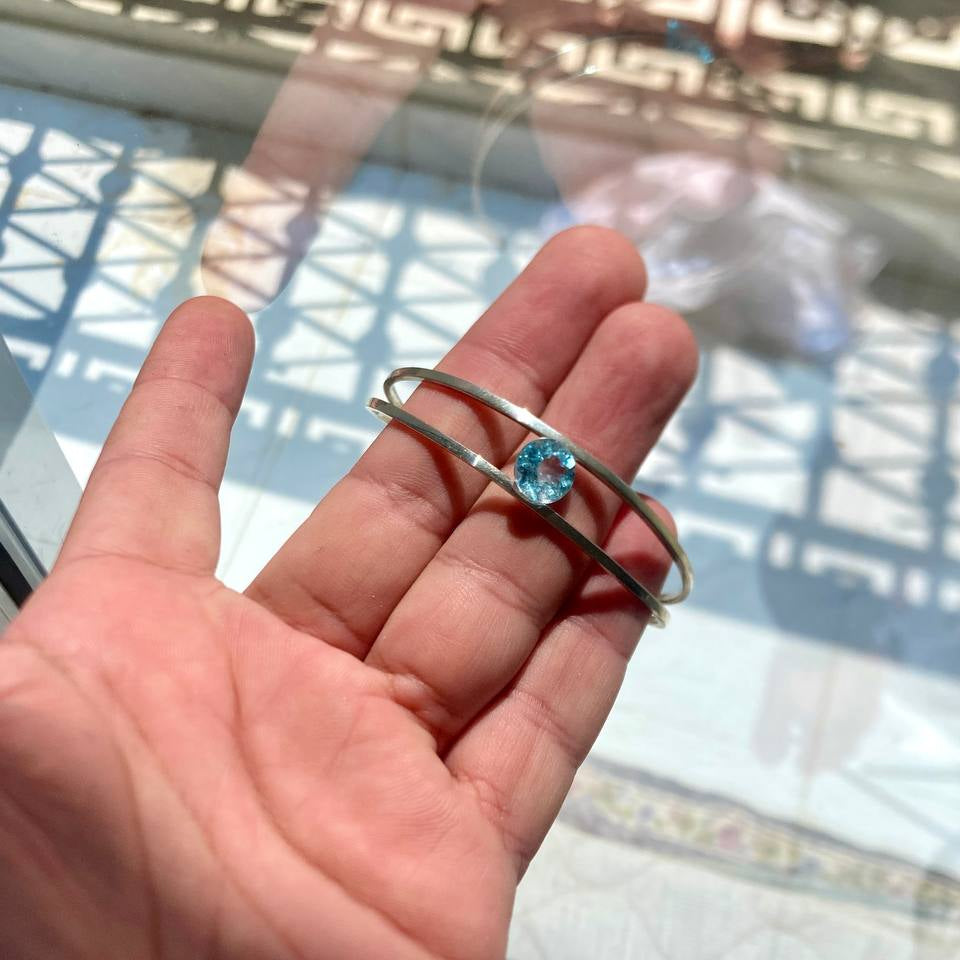 Hand holding a silver bracelet with a blue topaz gemstone in front of a reflective glass surface.