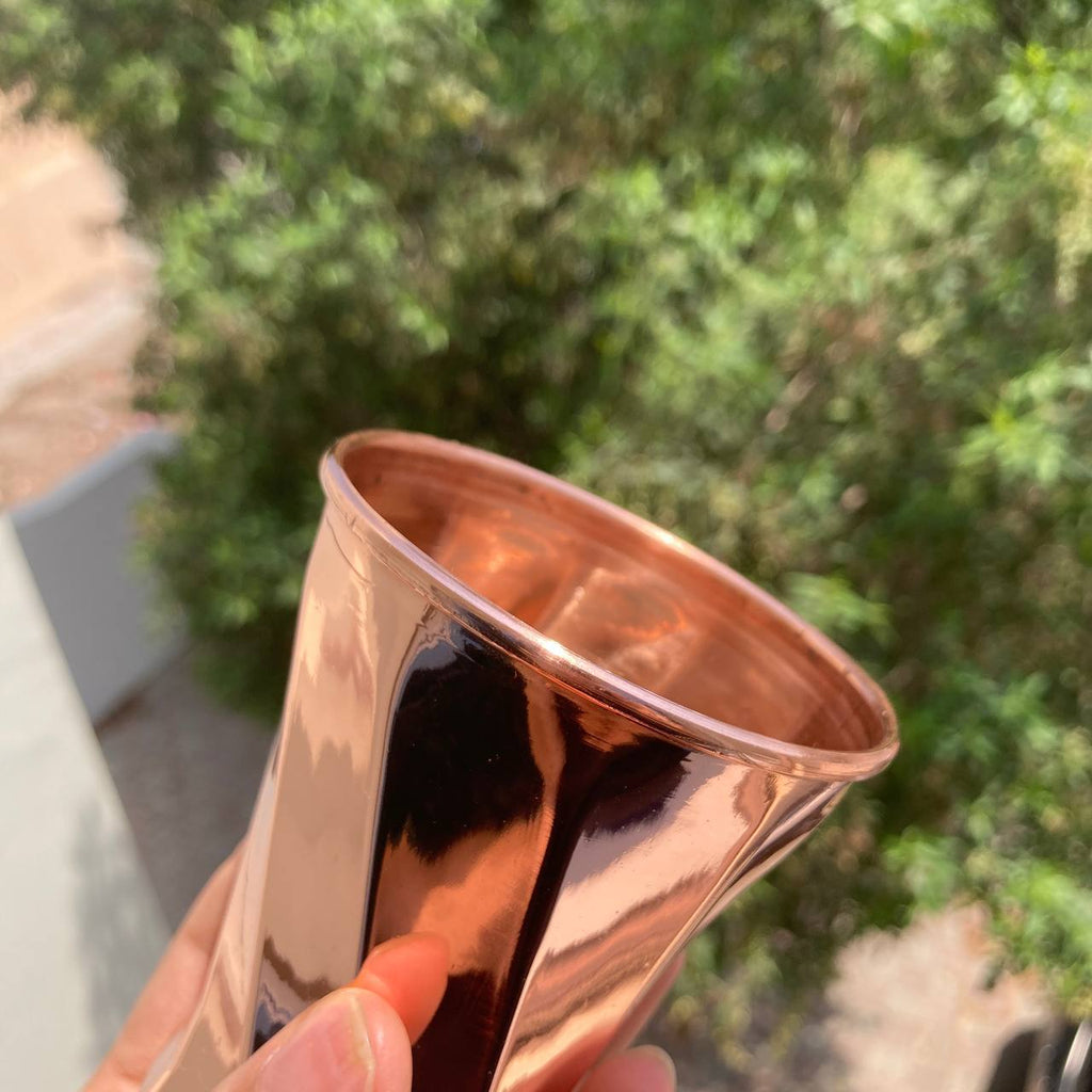 A close-up of a hand holding a shiny copper-colored metal tumbler outdoors, with blurred green foliage and a light-colored wall in the background.