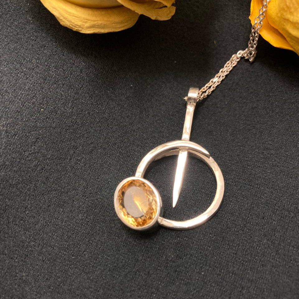 Silver pendant with a yellow gemstone on a dark surface with yellow flowers.