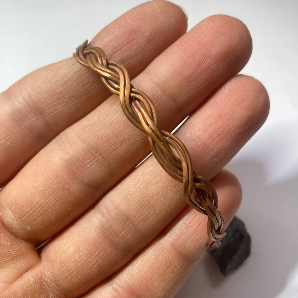 A close-up of a hand holding a thin braided metal bracelet or chain in a warm copper-gold tone against a plain light background.