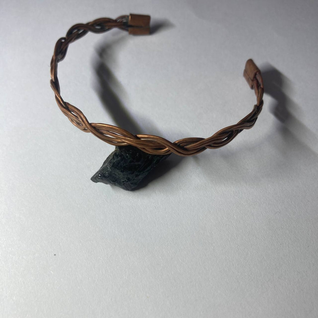 A twisted copper wire cuff bracelet with a small dark stone pendant, displayed on a light gray surface.