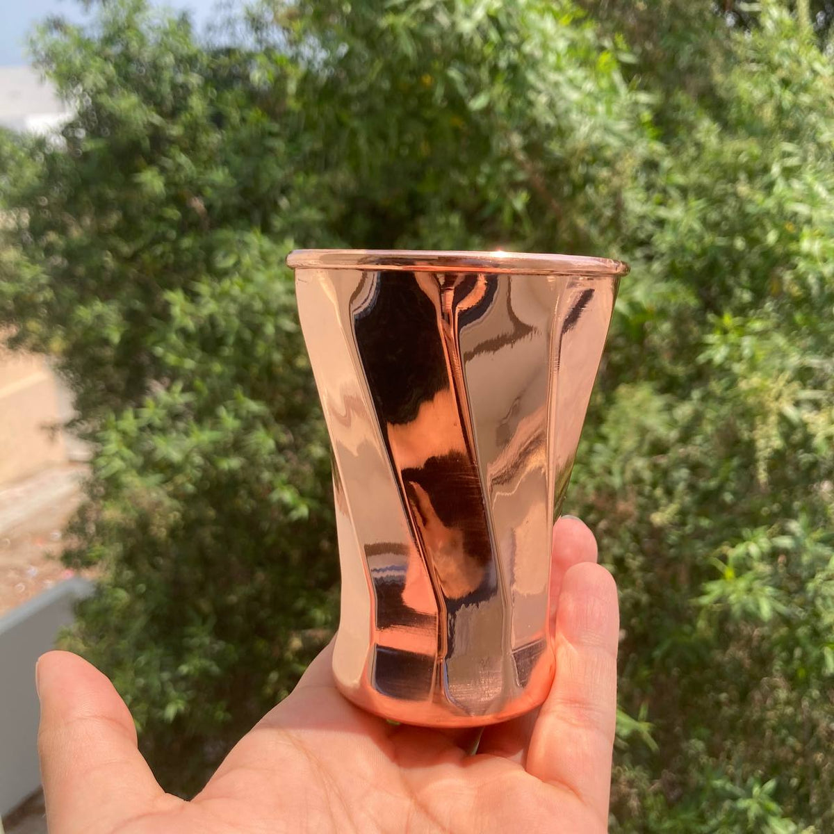 A hand holding a reflective copper-colored metal cup with a faceted, wavy design, set against a blurred background of green outdoor foliage.