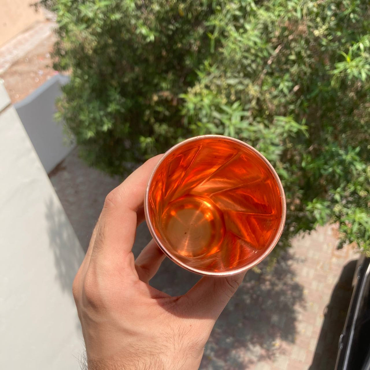 A hand holding a small copper-colored glass filled with amber liquid, photographed outdoors with green foliage and a light-colored wall in the background.
