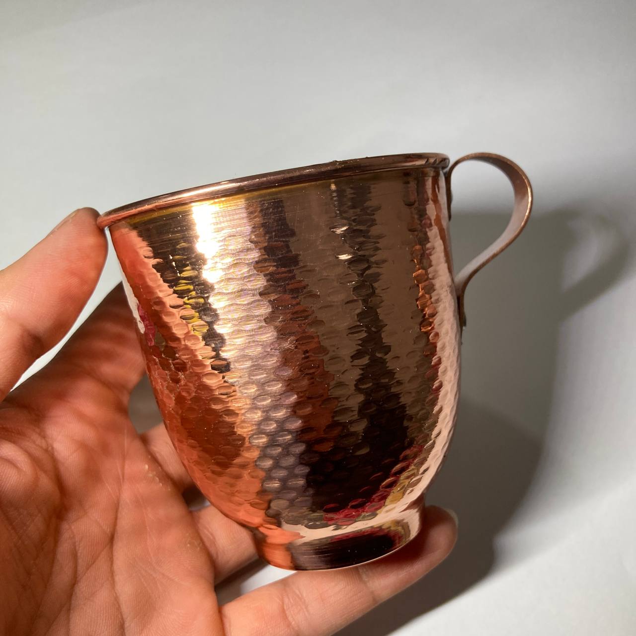 A hand holding a hammered copper mug with a shiny metallic finish against a plain light background.