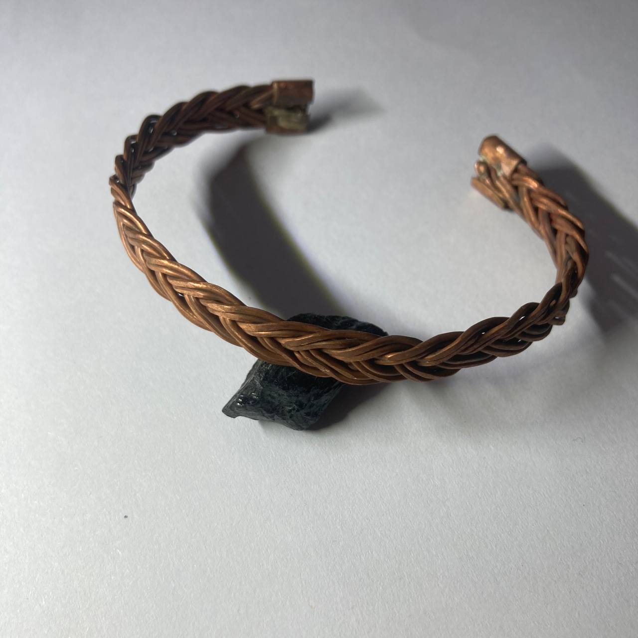 A handmade braided copper cuff bracelet with a dark stone accent, photographed on a neutral light gray background.