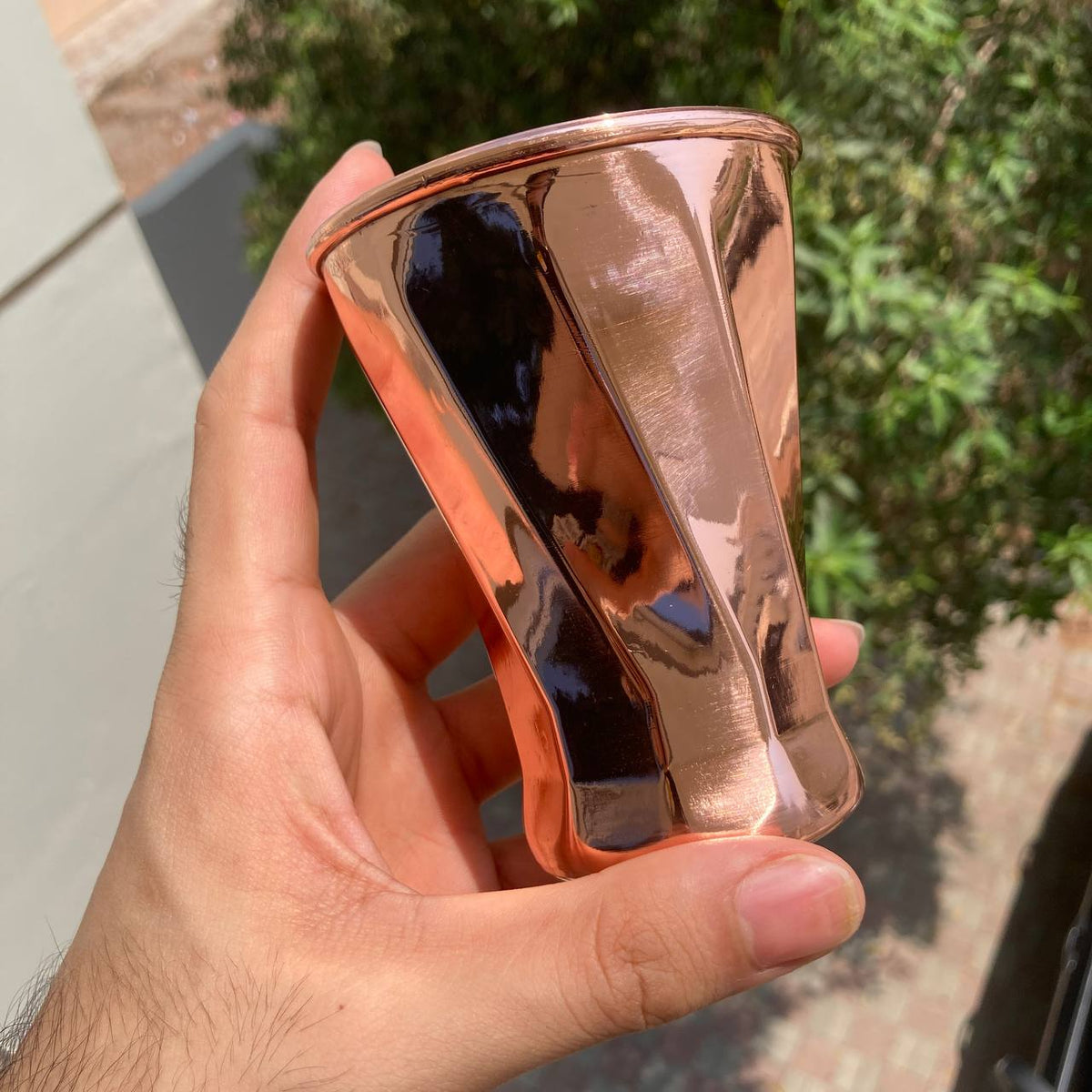A hand holding a small, faceted copper-colored metal tumbler outdoors with green foliage and a building wall in the background.