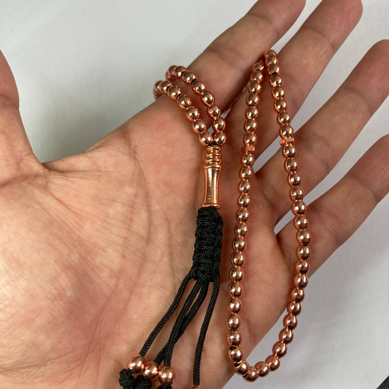copper beaded rosary with a black braided tassel, held in a person's hand against a plain light background.