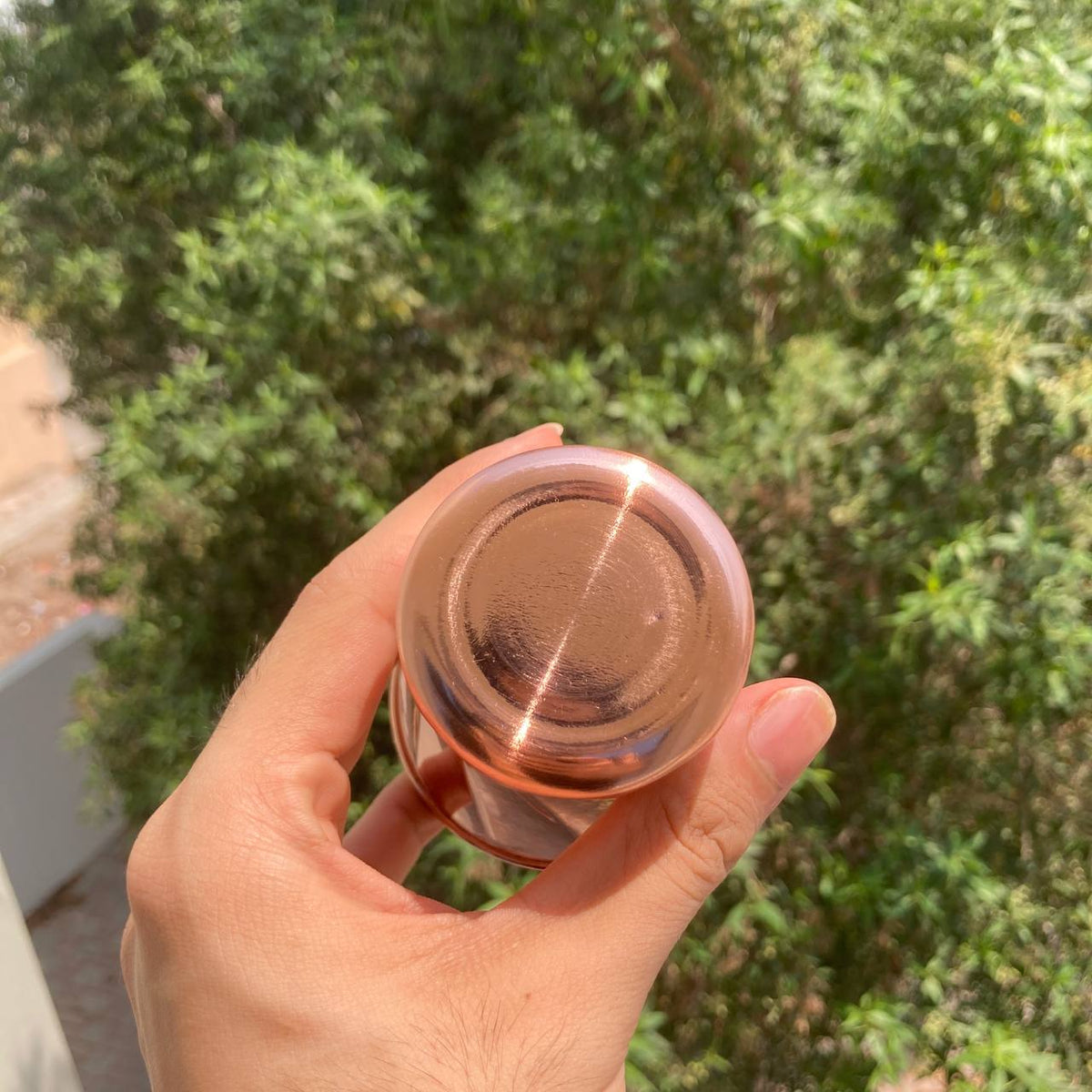 A hand holding a small, round, metallic copper cosmetic container against a blurred background of green foliage.