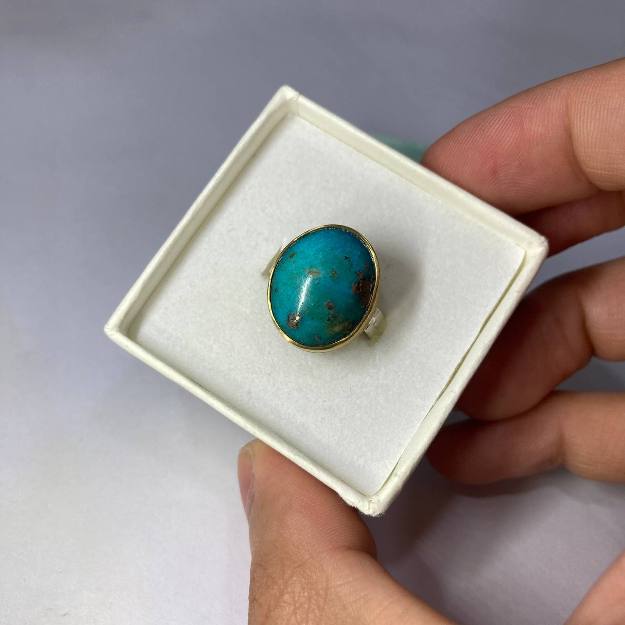 Turquoise ring in a white box held by a hand against a gray background