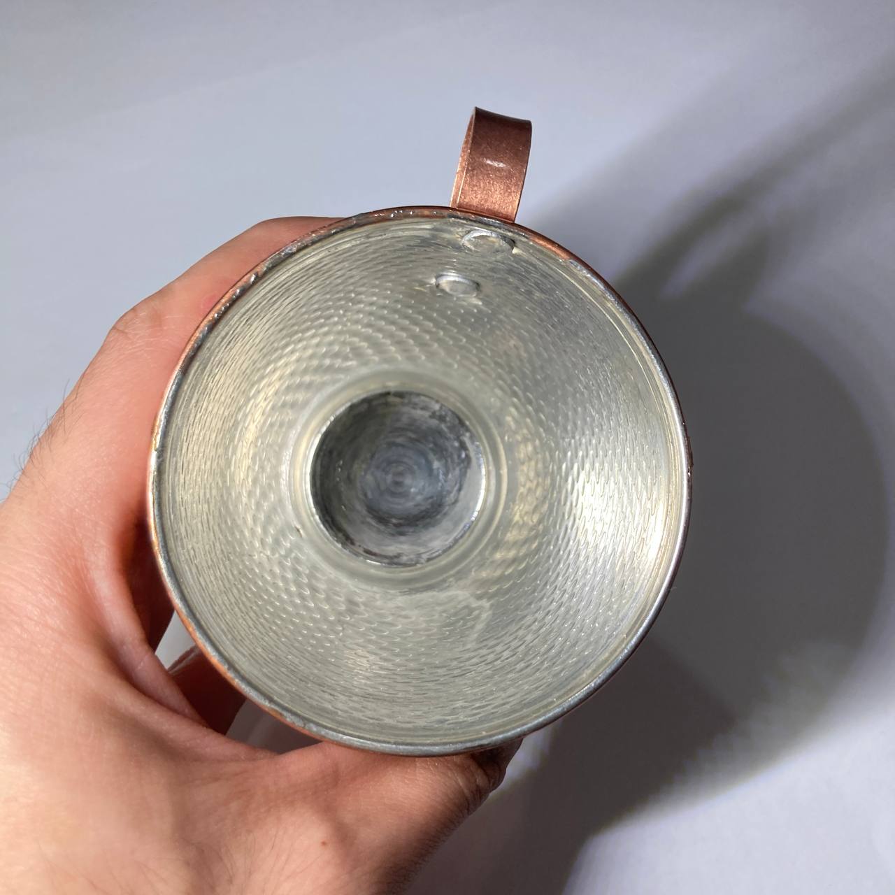 A hand holding a copper mug, showing the textured silver-toned interior and copper-colored exterior and handle against a plain light background.