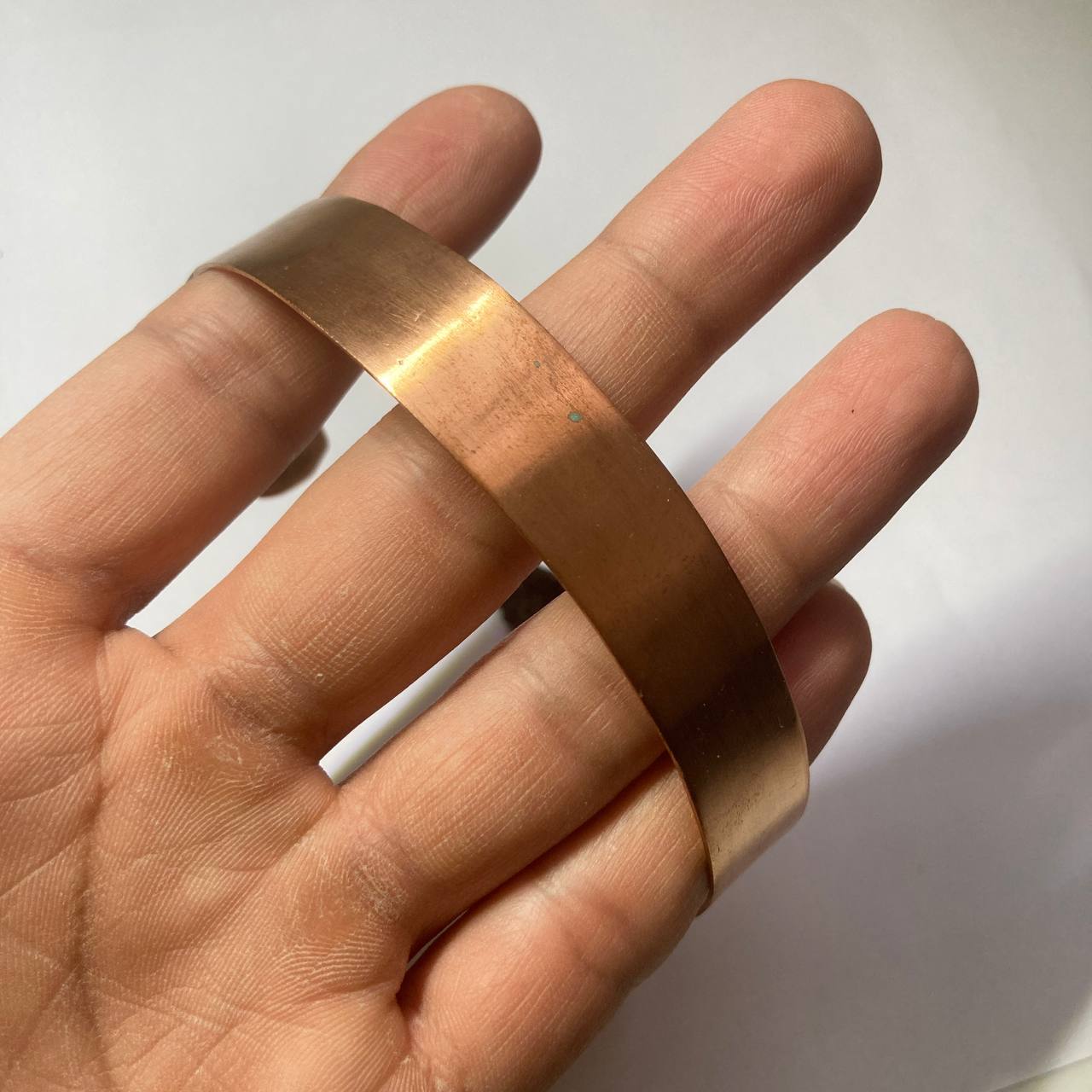 A wide, polished copper-colored metal cuff bracelet being held on a person's hand against a plain light background.