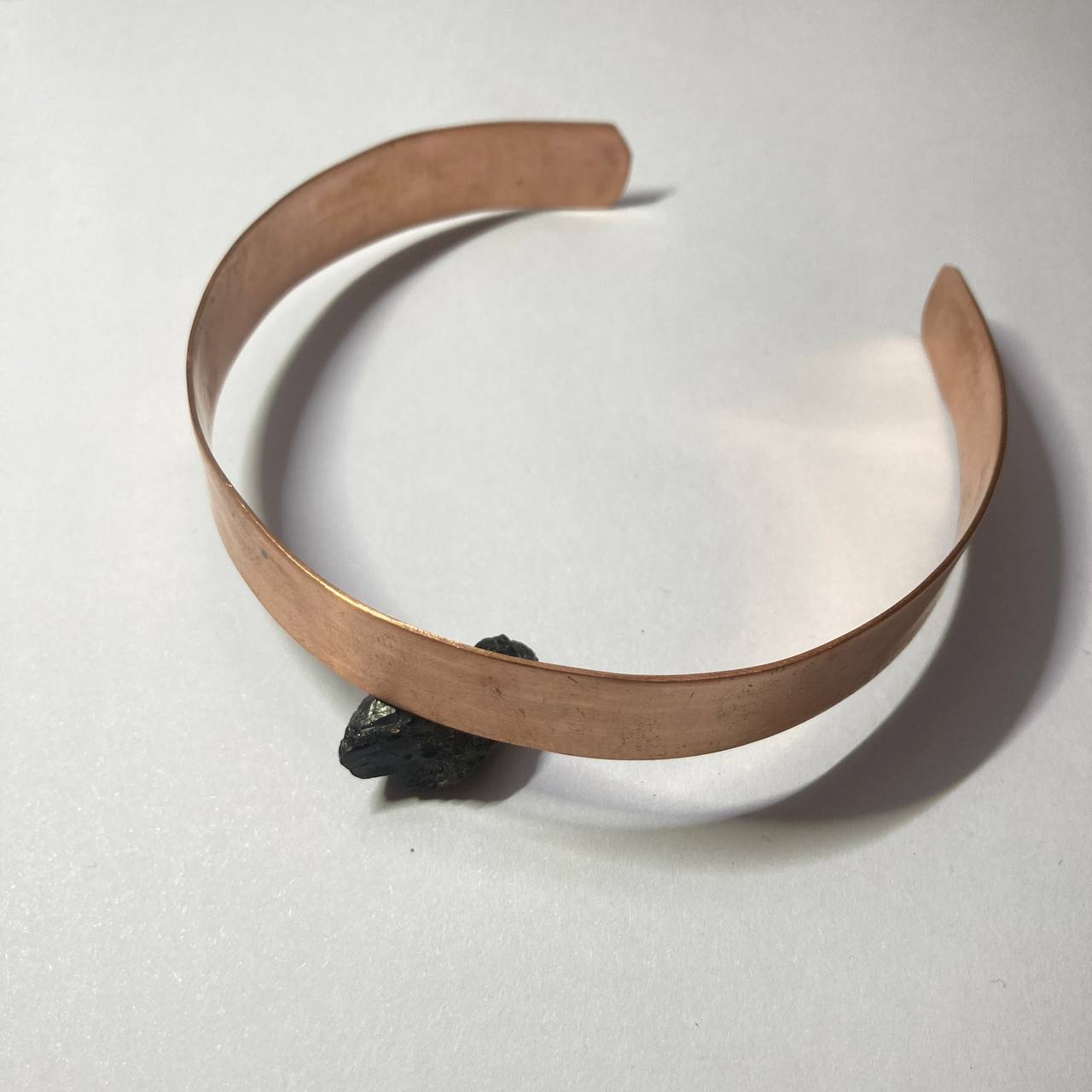 A handmade copper cuff bracelet with a polished, hammered finish, resting on a light gray surface with a small black stone prop.