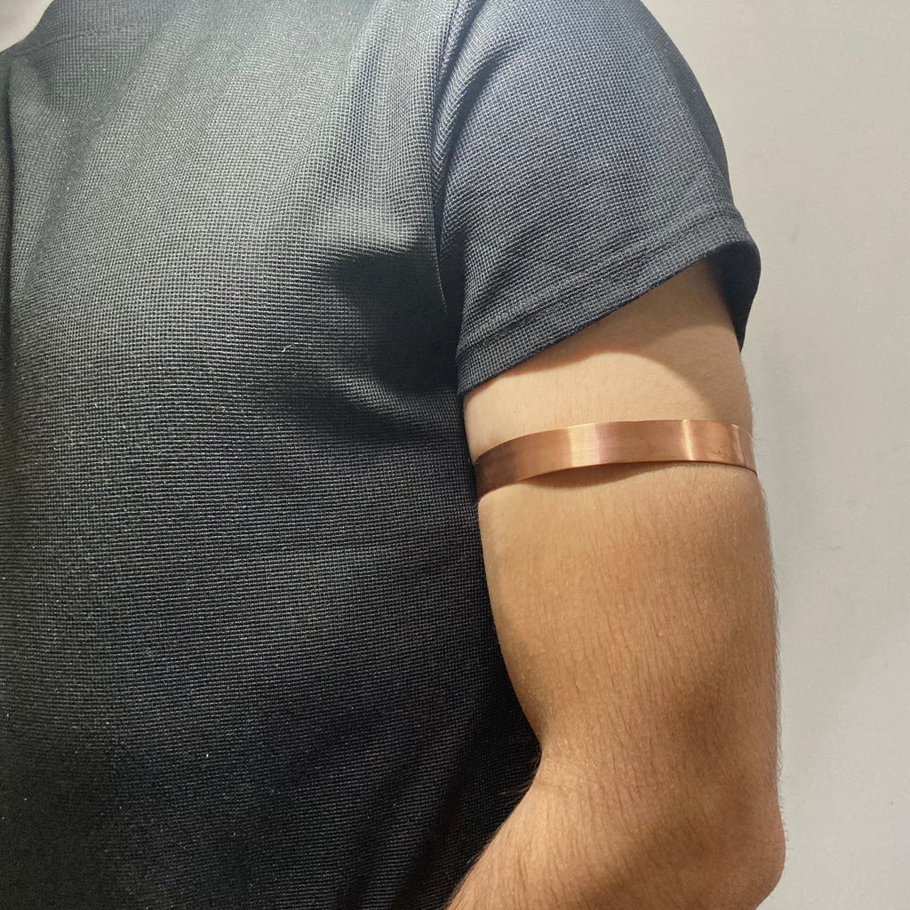 A close-up of a person's upper arm wearing a thin, polished copper-colored metallic band, shown against a dark gray t-shirt and a plain light background.