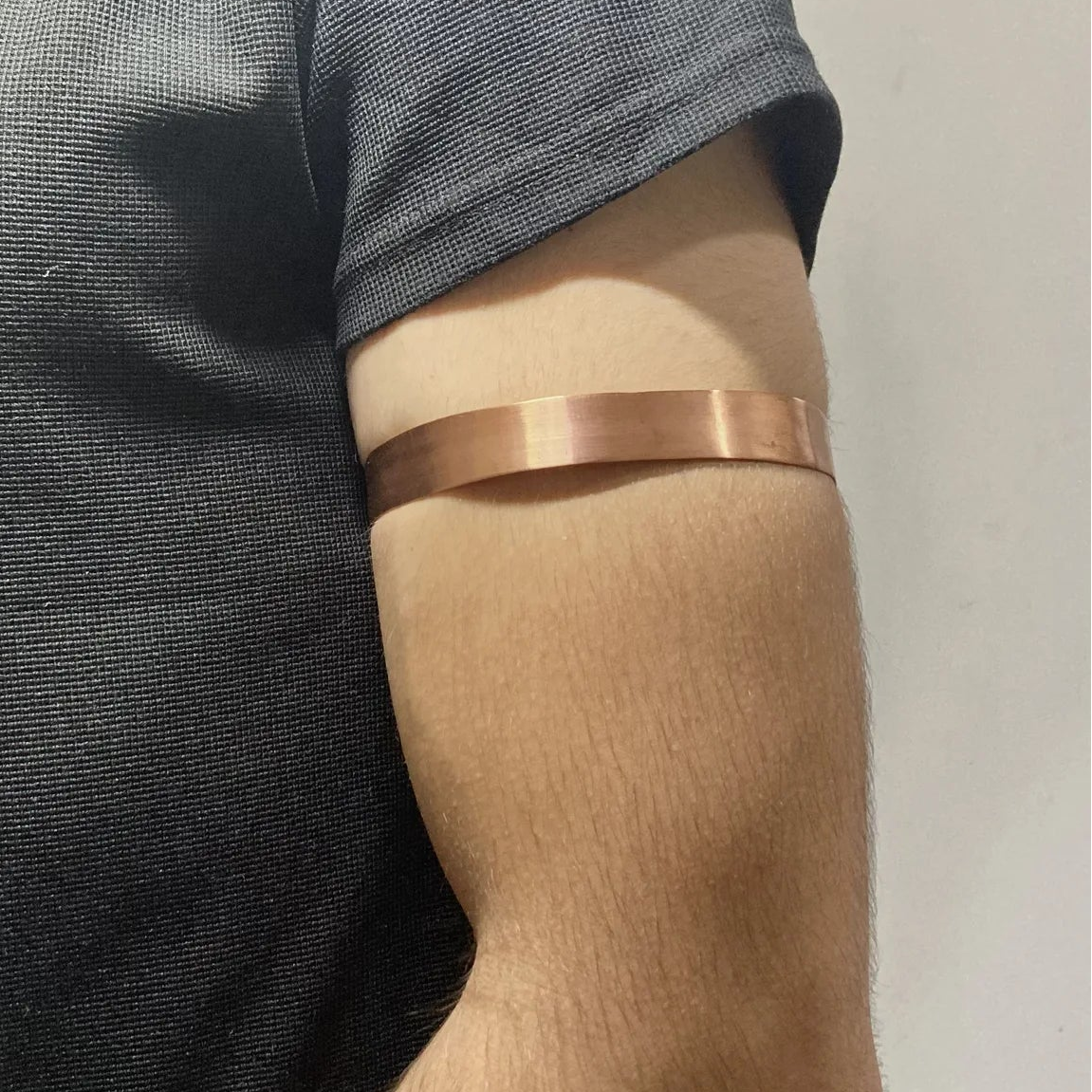 A close-up of a person's upper arm wearing a thin, polished copper-colored metallic band, shown against a dark gray t-shirt and a plain light background.