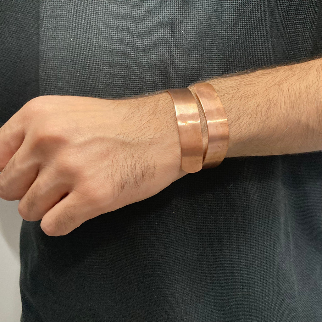 A close-up of a person's wrist wearing two thick, polished copper-colored metal cuff bracelets against a dark textured shirt.
