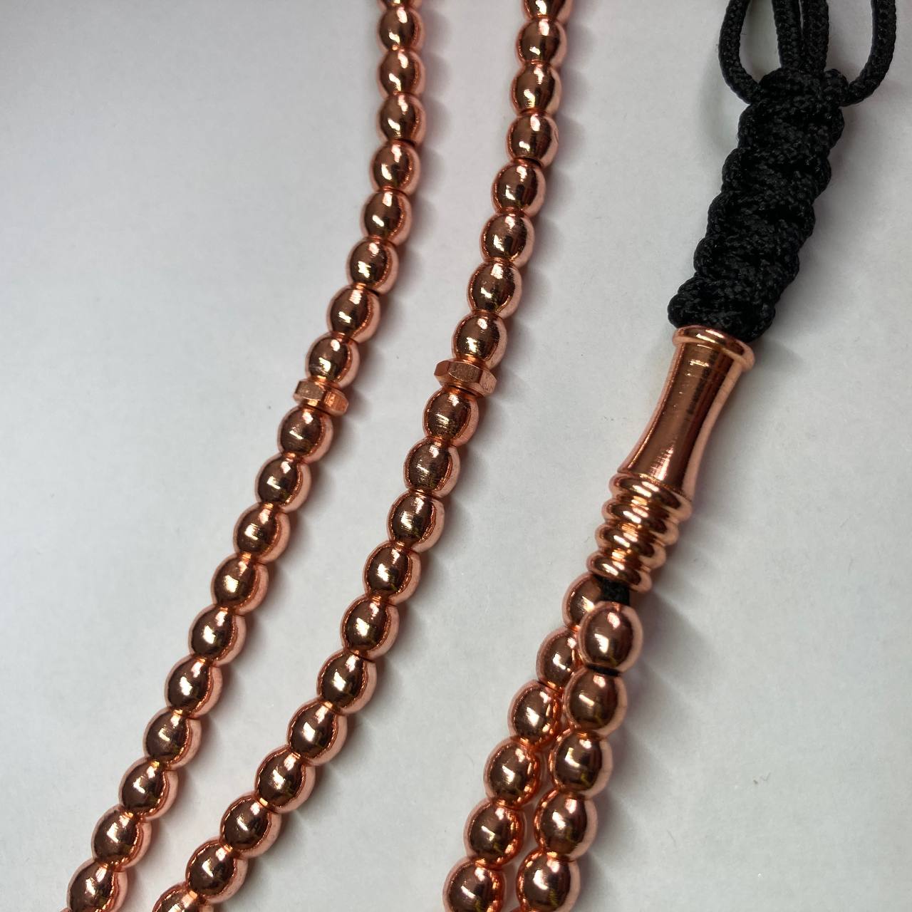 A close-up of a metallic copper beaded rosary or misbah or tasbih accessory with a black braided cord loop on a plain light background.