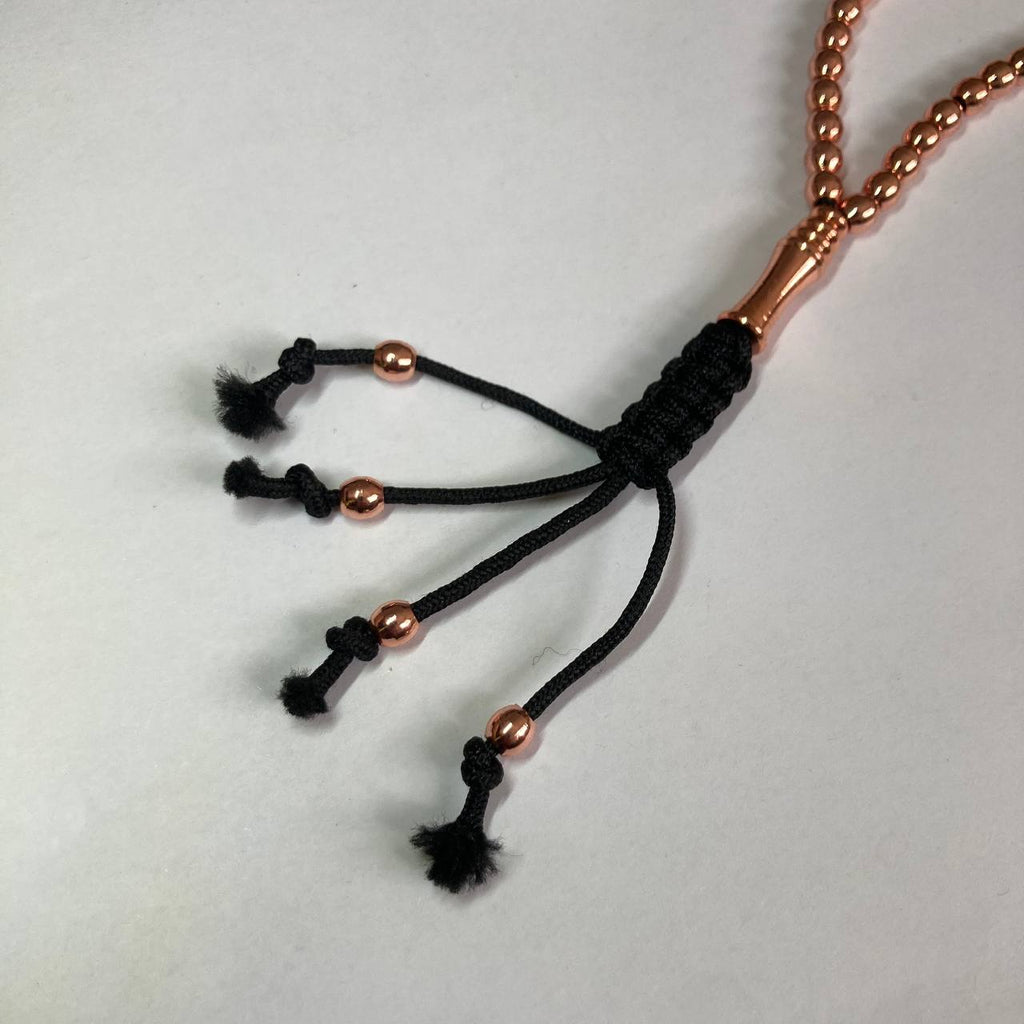 A close-up of a rosary/misbah/tasbih head with a black braided cord tassel design and copper metallic beads, photographed on a plain light background.