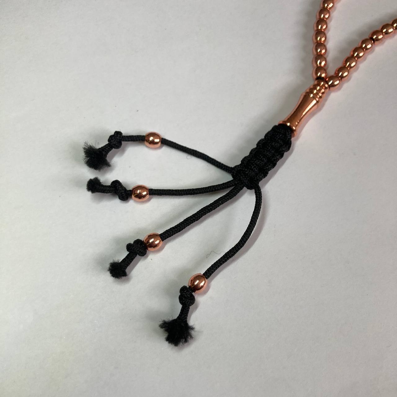 A close-up of a rosary/misbah/tasbih head with a black braided cord tassel design and copper metallic beads, photographed on a plain light background.
