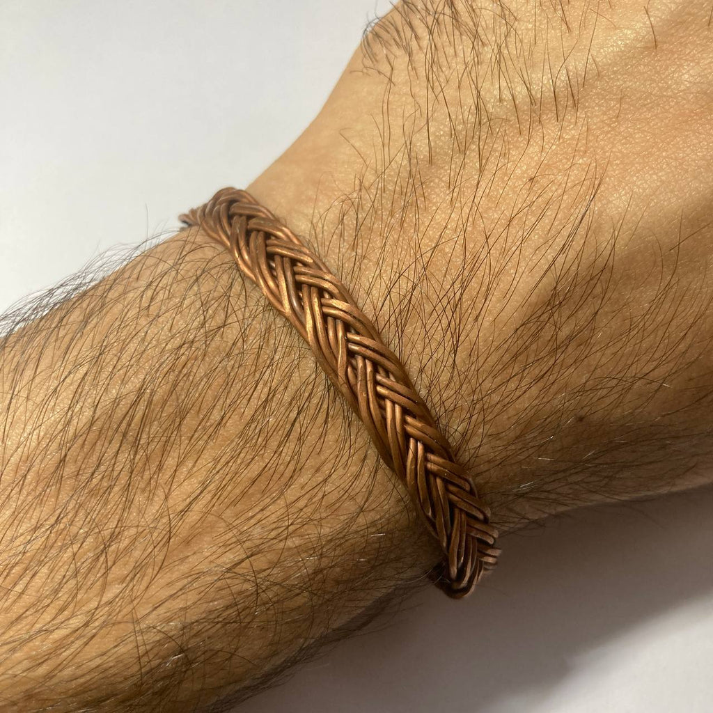 A close-up of a braided copper bracelet worn on a hairy wrist, showing the metallic woven pattern against the skin.