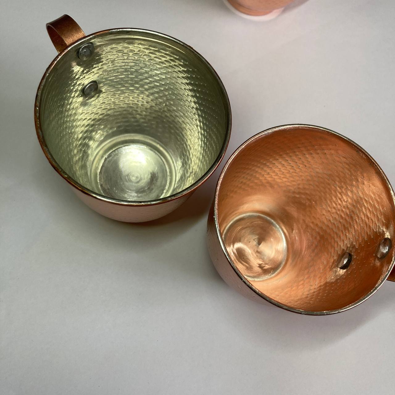 Two hammered copper mugs with copper-colored interiors and silver-toned rims, shown on a light gray surface.